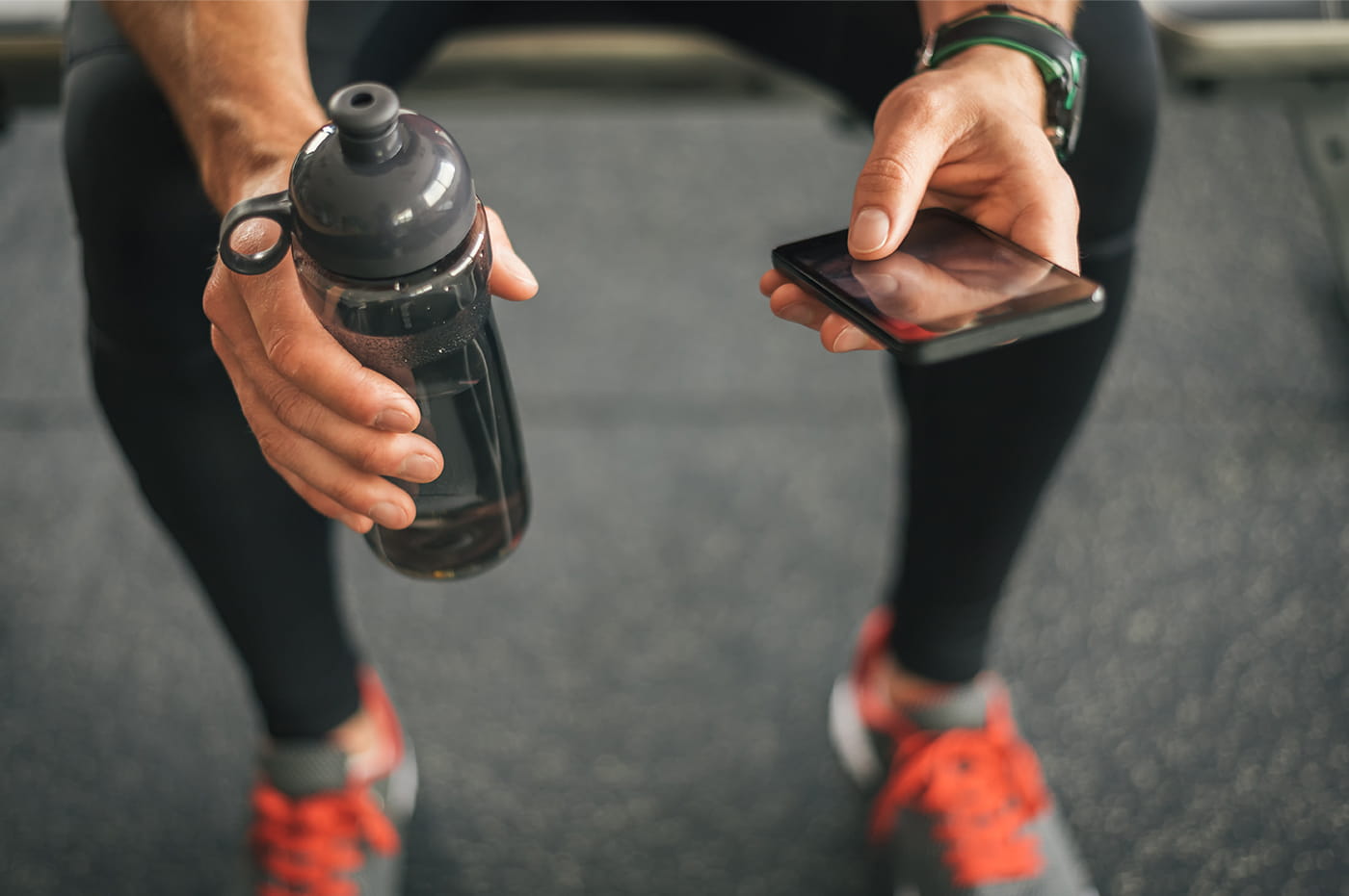 A man's hands holding a water bottle and texting on phone, wearing bright athletic shoes.