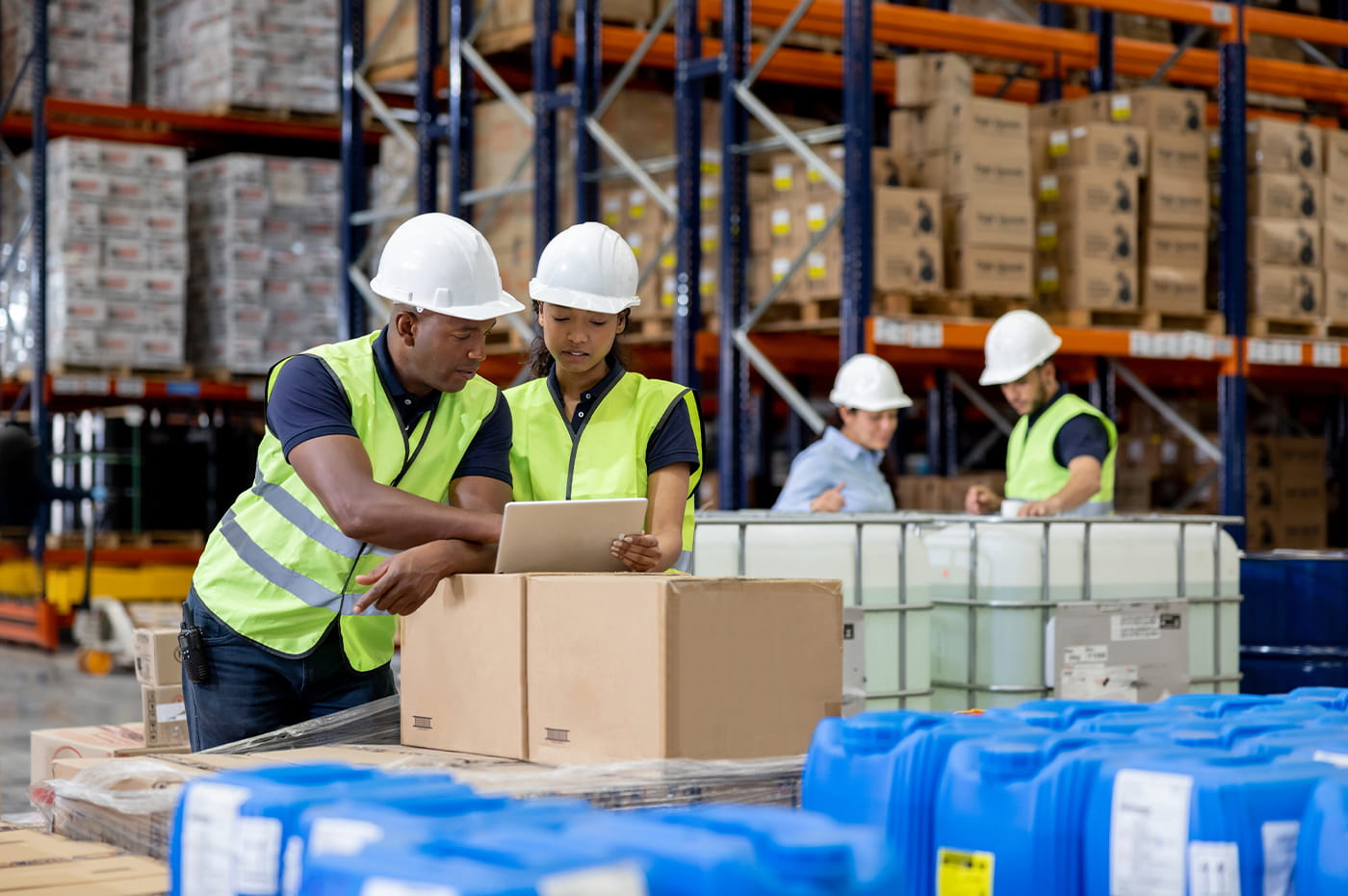 Group of workers in a warehouse looking at a tablet, wearing construction vests and hard hats